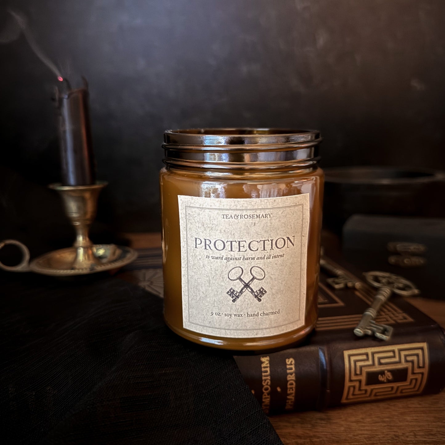 A candle in a clear glass jar with a silver metal lid and a cream label reading 'PROTECTION' styled on a dark tabletop with an old book, a key, and a candle holder in the background.