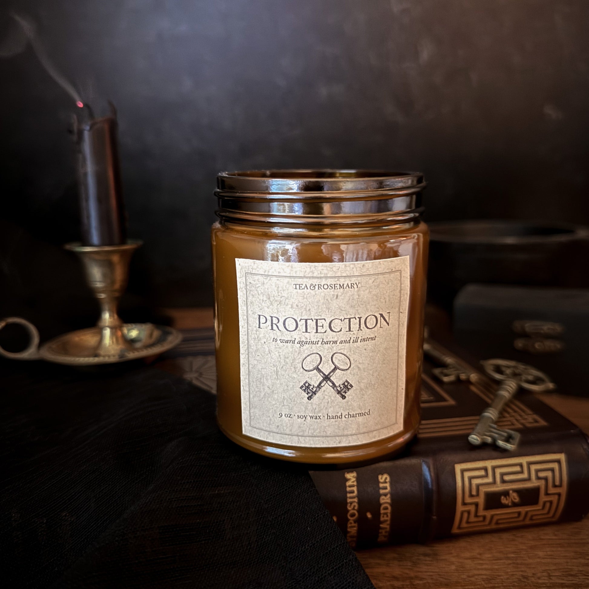 A candle in a clear glass jar with a silver metal lid and a cream label reading 'PROTECTION' styled on a dark tabletop with an old book, a key, and a candle holder in the background.