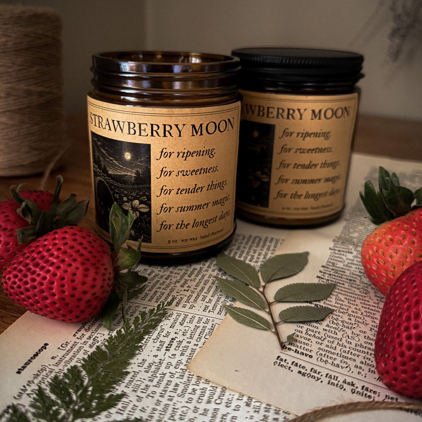 Two Strawberry Moon candles on a surface with strawberries and leaves.