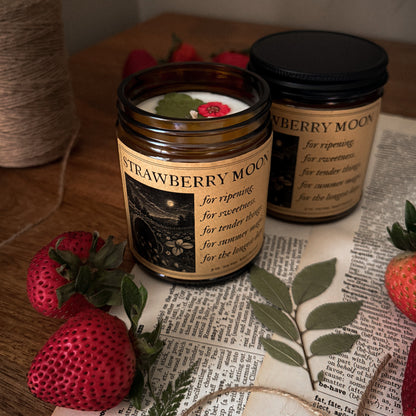 Two Strawberry Moon candles on a surface with strawberries and newspaper.